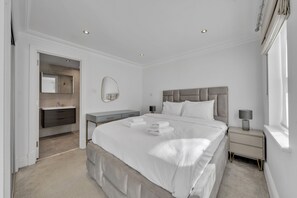 A modern bedroom with a large bed, bedside tables, a nightstand, a mirror, and a bathroom visible through an open door. - Luxury 3-bedroom Apartment Next to Harrods (London)