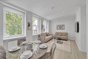A dining area with a table set for four, a sofa, and a television in a room with large windows. - Luxury 3-bedroom Apartment Next to Harrods (London)