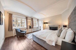 Ein Schlafzimmer mit einem großen Bett, einem Sessel, einem Kamin und Blick auf ein Gebäude durch die Fenster.