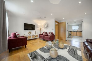 A modern living room with a flat-screen TV, red sofas, a coffee table with decorative items, and a kitchen visible through an open door. - Three Bedroom Apartment Next to the Station (London)