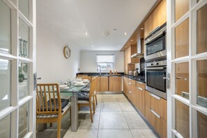 A modern kitchen with wooden cabinets, stainless steel appliances, and a glass dining table. - Three Bedroom Apartment Next to the Station (London)
