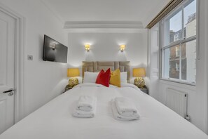 Ein Hotelzimmer mit einem großen Bett, zwei Nachttischlampen, einem Flachbildfernseher und einem Fenster mit Blick auf Gebäude.