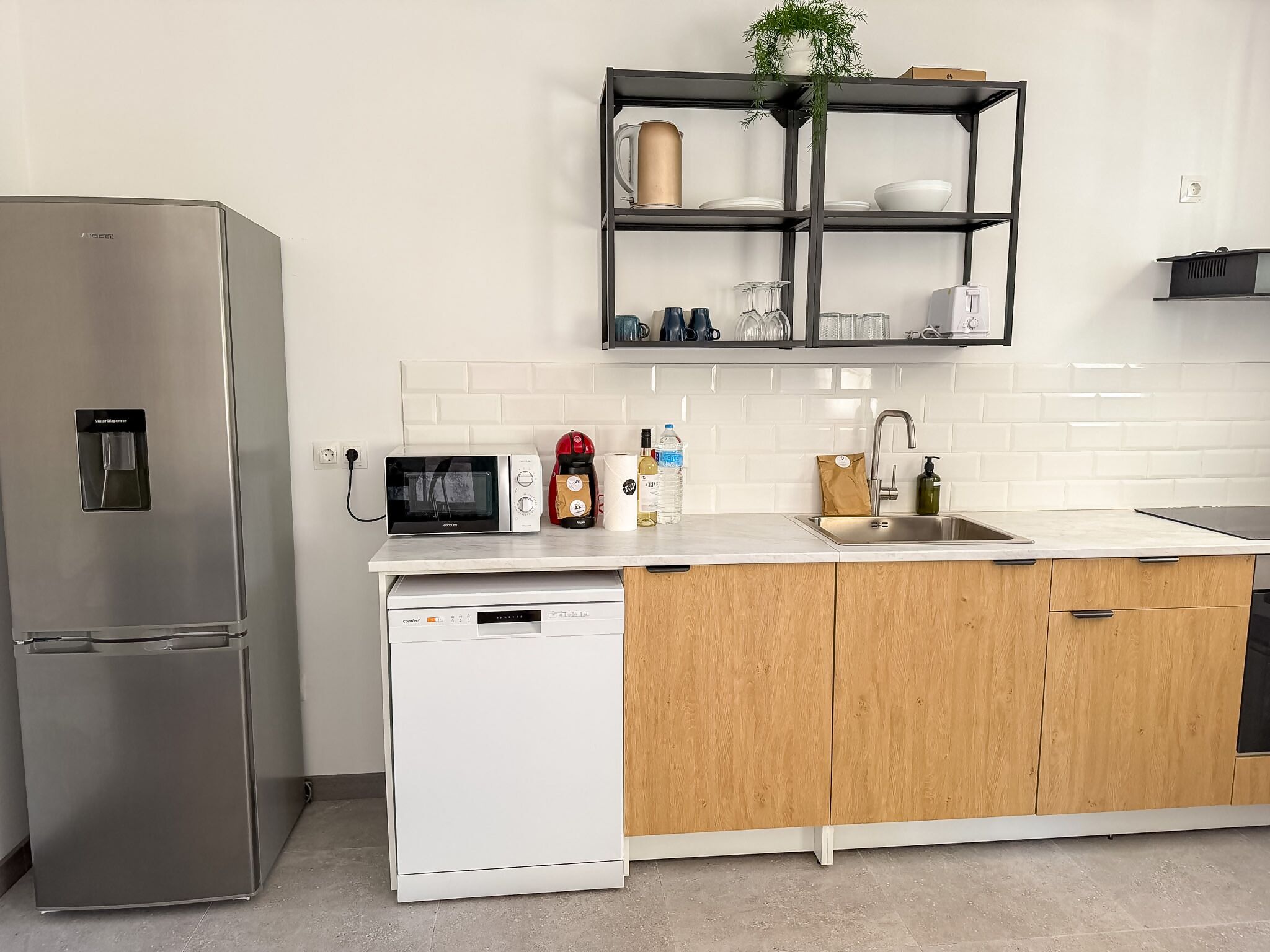 A modern kitchen with a stainless steel refrigerator, a dishwasher, wooden cabinets, a white countertop, a black shelf with glassware, and a microwave.