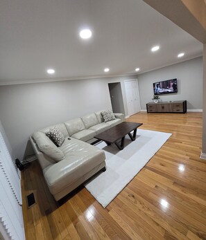 A modern living room with a sectional sofa, a coffee table, wooden flooring, recessed ceiling lights, and a television mounted on the wall. - Delightful 2-bedroom apartment with balcony in Sterling Heights (Sterling Heights)