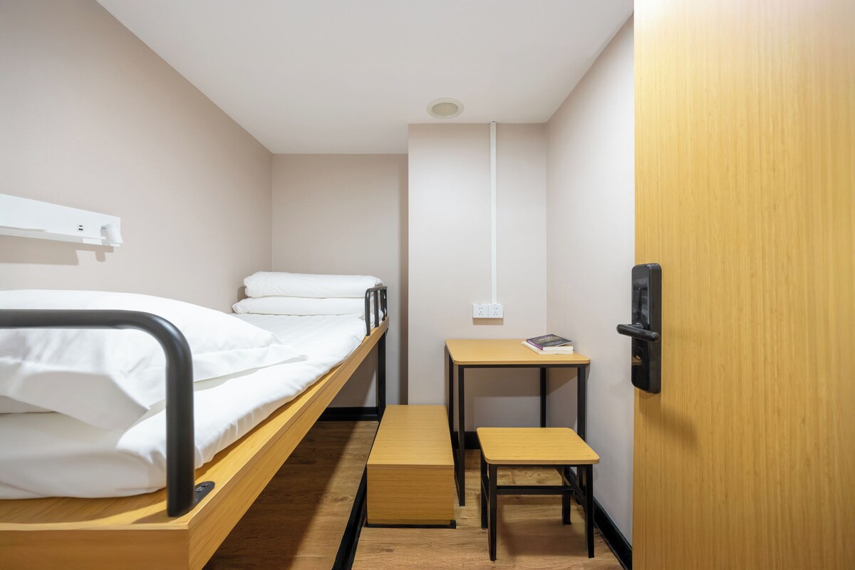 Una cama individual con estructura de madera, una mesita de madera, un banco y una puerta con manija.