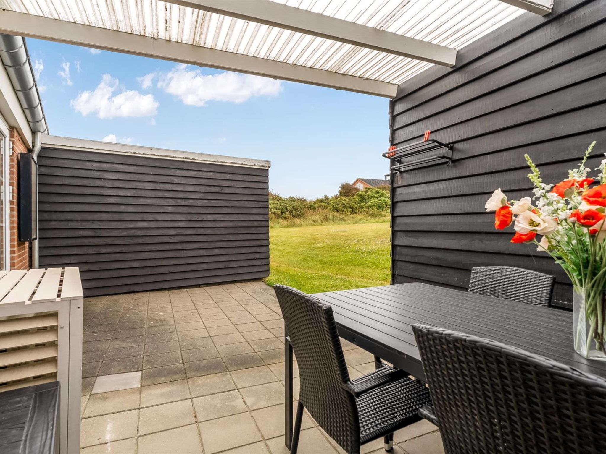 An outdoor patio with a table and chairs, a covered area with a metal roof, and a garden view.