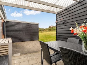 Um pátio externo com mesa e cadeiras, uma área coberta com telhado metálico e vista para o jardim.