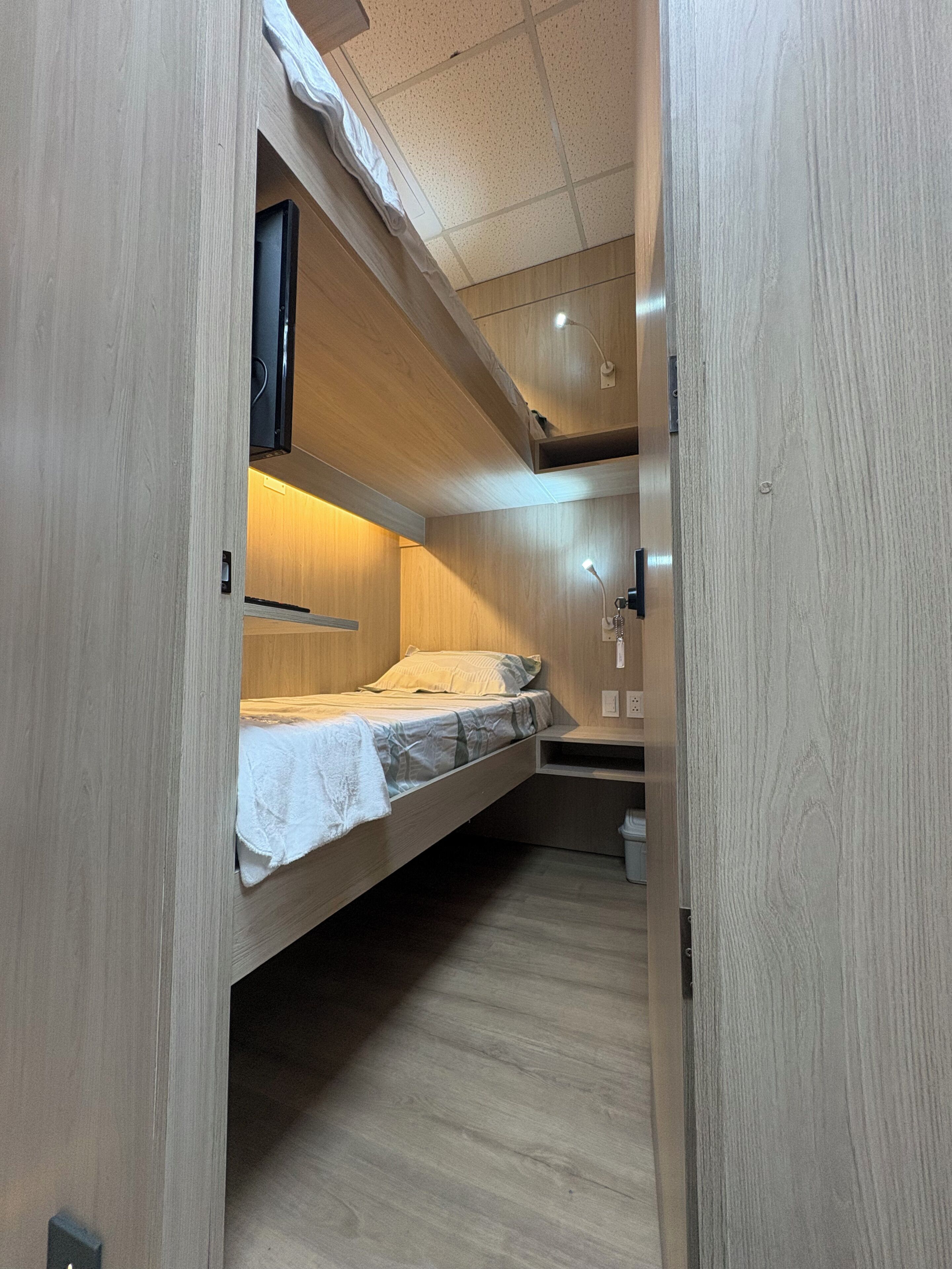 A compact, single-bed room with a built-in bench and a small shelf, featuring a wooden finish and a ceiling with recessed lighting.