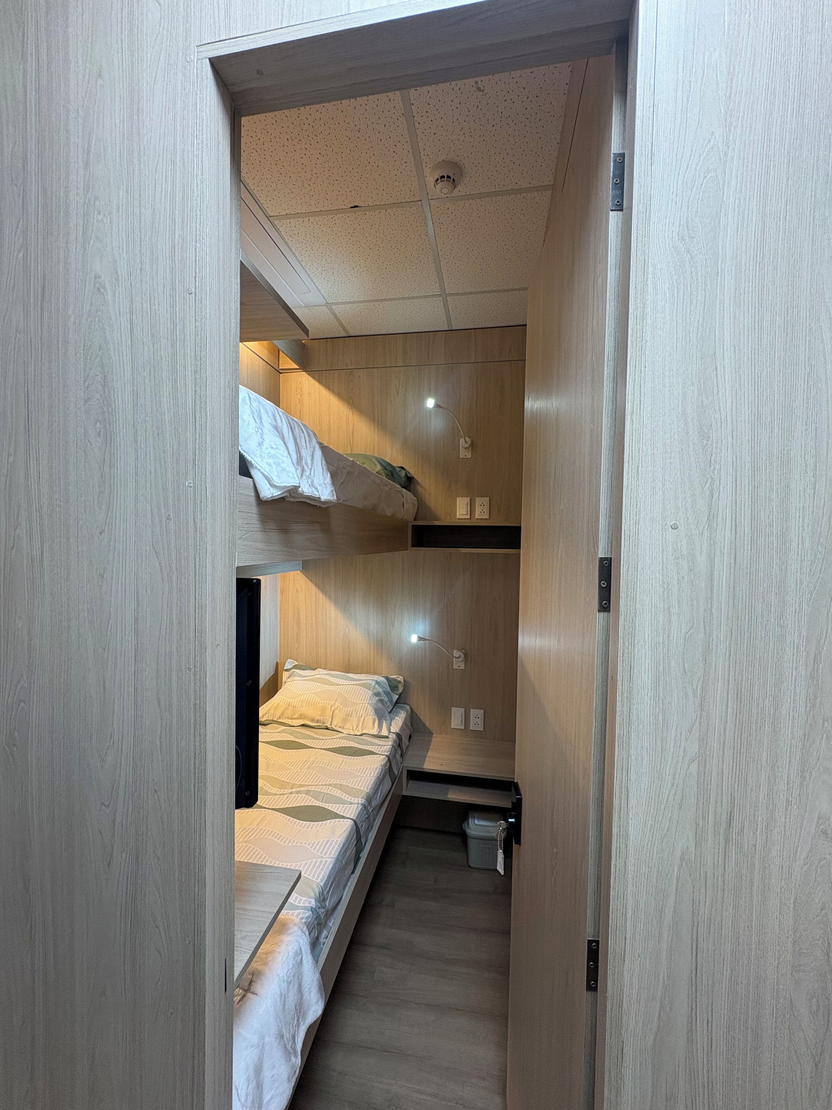 A narrow hallway leading to a bunk bed room with a wooden floor and a ceiling with a smoke detector.