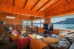 A cabin interior with a wooden ceiling, a dining area, a television, and a view of a lake. - Rotoiti Boathouse - Waterfront, Jetty, Kayaks (Rotoiti Forest)