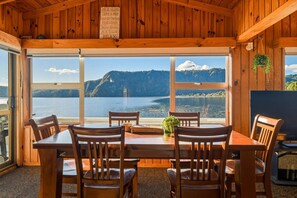 A wooden cabin with a dining table and chairs, large windows with a view of a lake and mountains, and a sign with relaxation activities. - Rotoiti Boathouse - Waterfront, Jetty, Kayaks (Rotoiti Forest)