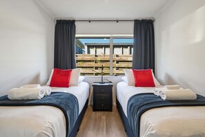 Ein Hotelzimmer mit zwei Betten, einem Nachttisch und einem Fenster mit Blick auf eine Holzterrasse.