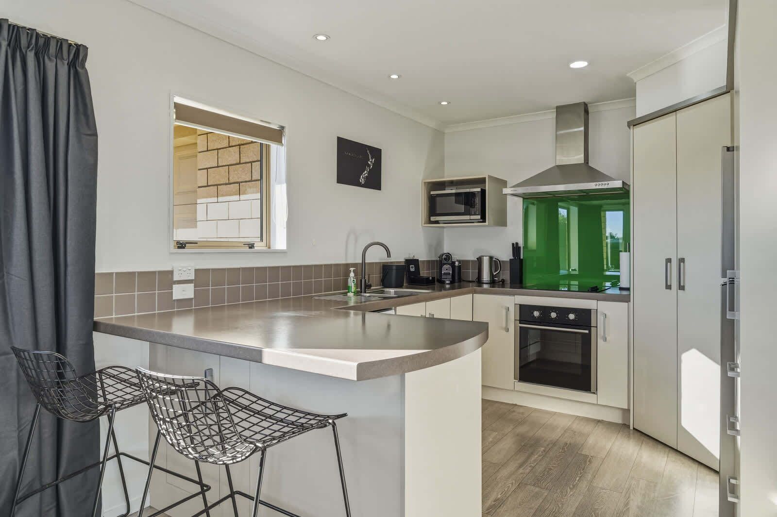 A modern kitchen with a central island, a microwave, a stove, and a green-tinted oven. There are bar stools and a window with a view.