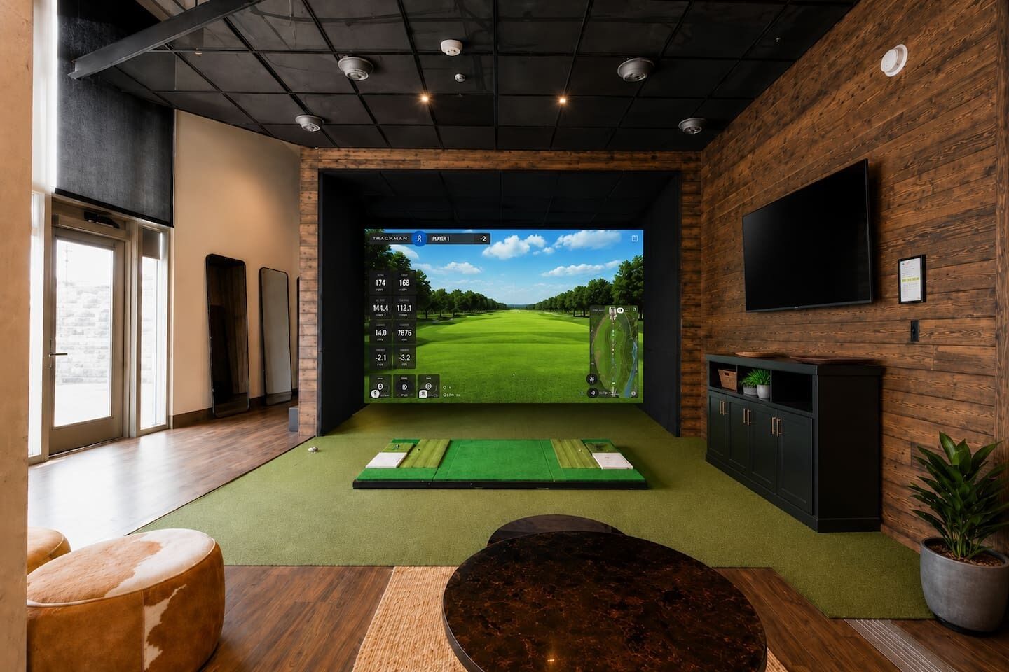 A modern room with a large screen displaying a golf game, a green practice mat, a wooden cabinet, and a potted plant.