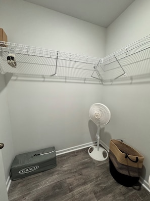 Ein kleiner Raum mit einem Metallregal, einem Standventilator, einer Aufbewahrungsbox und einem gewebten Korb.