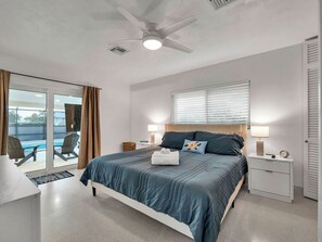 Ein Schlafzimmer mit Bett, Deckenventilator, Fenster mit Jalousien, Nachttisch mit Lampe und Blick auf einen Pool.