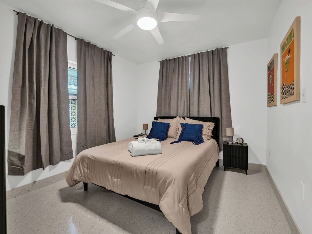 A bedroom with a bed, curtains, a ceiling fan, a nightstand, and wall art.