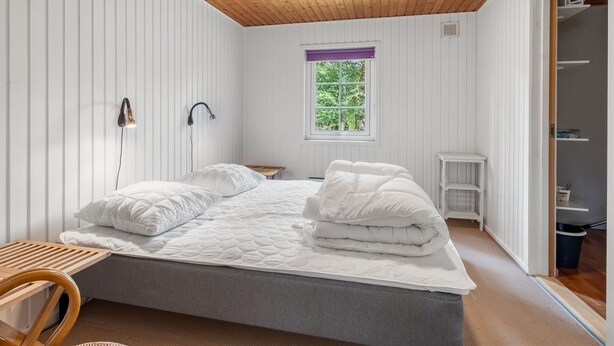 Ein Schlafzimmer mit Holzboden, einem Bett mit weißer Bettwäsche, einem Fenster mit Blick auf Grünflächen und einem kleinen Nachttisch.
