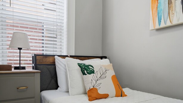 A neatly made bed with white bedding, a grey nightstand with a lamp, and a wall with a painting. - Bright Upper-Level 3BR Near WashU & Forest Park (St. Louis)