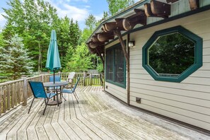 Une terrasse en bois avec une table et des chaises, un parasol vert et un bâtiment de style cabane doté d’une grande fenêtre.