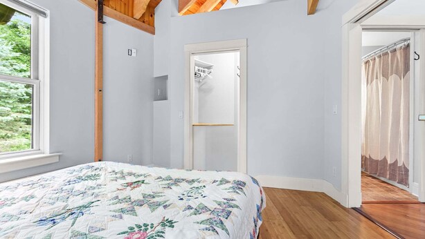 Ein Schlafzimmer mit einem Bett, Holzbalken, einer Tür und Blick durch ein Fenster.