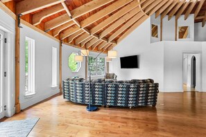 A spacious room with a high ceiling, wooden beams, a round seating area, a flat-screen TV, and a door leading to another room. - Cranberry Cottage - NEW! Home in Trenton (Trenton)