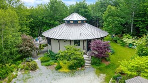 Une structure de type belvédère, dotée d’un toit conique, entourée d’une végétation luxuriante et accessible par un sentier de gravier.