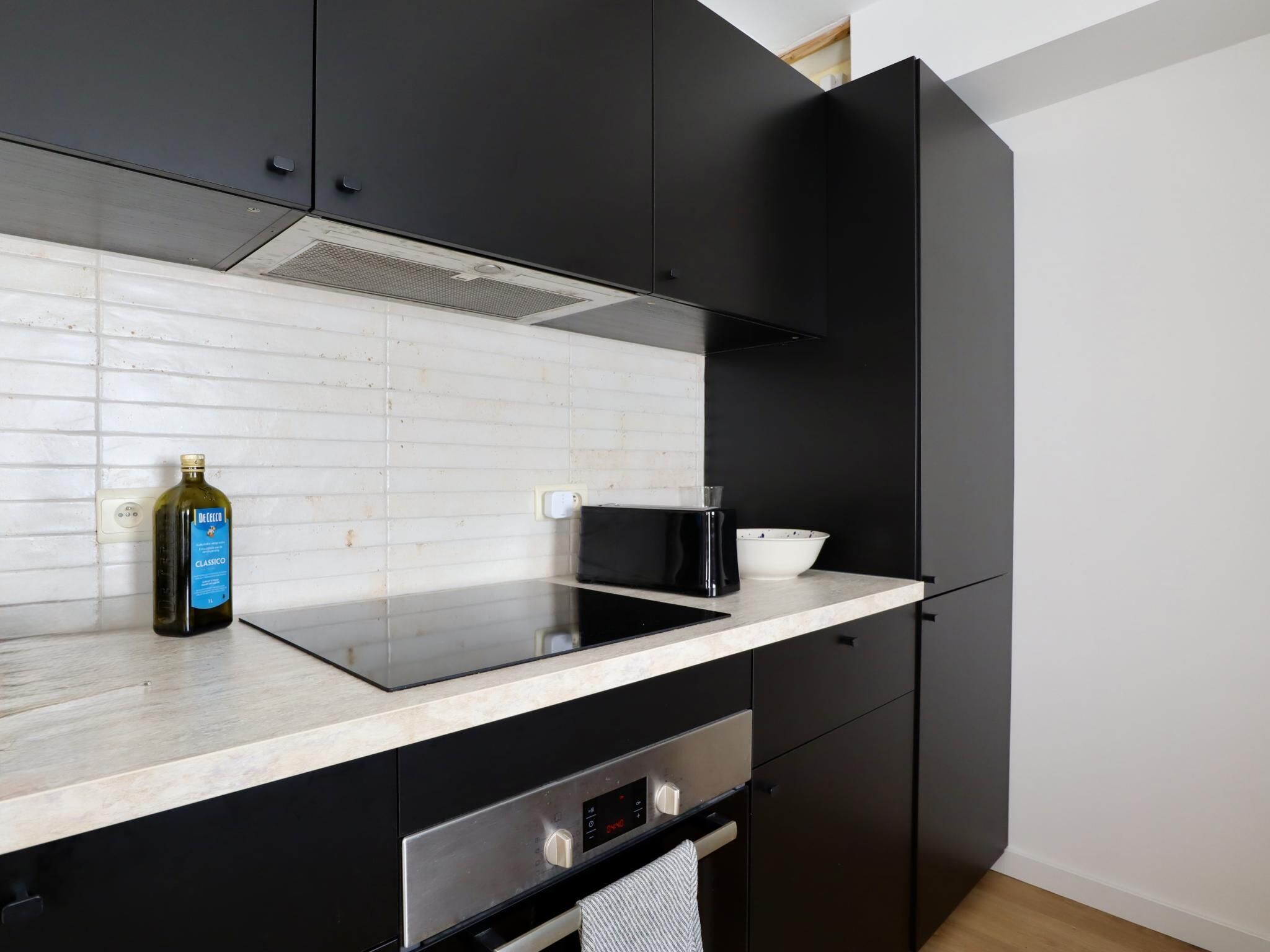 A modern kitchen with black cabinets, a white tiled backsplash, a stainless steel range hood, a black induction cooktop, a white bowl, and a bottle of olive oil.