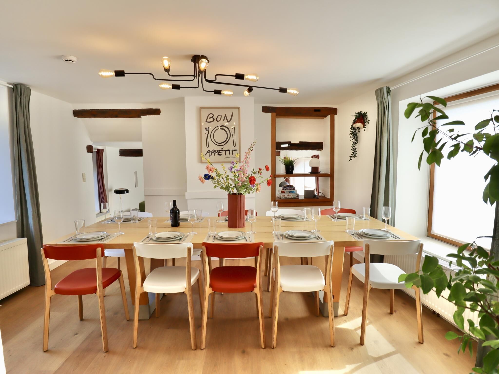 Un coin repas avec une table en bois dressée pour six personnes, entourée de chaises dotées de coussins rouges et blancs. Un vase rempli de fleurs est placé au centre.