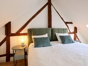 Ein Schlafzimmer mit hoher Decke und freiliegenden Holzbalken, einem Bett mit weißer Bettwäsche und zwei grünen Kissen, sowie einem kleinen Nachttisch mit einer Lampe.