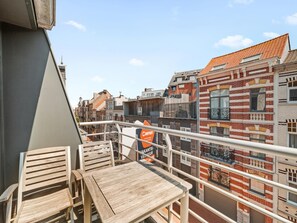 A balcony with wooden furniture and a view of a cityscape with buildings and a sign for a business. - Beautiful 2-Bedroom Apartment in City Center (Blankenberge)