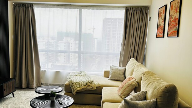 A modern living room with a sofa, two round coffee tables, a large window with curtains, and a city view. - 14th Floor 2B with Pool, Gym & King Bed in Lavington (Nairobi)