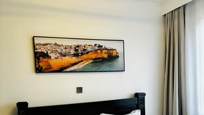A hotel room with a bed, a painting of a coastal town, and a window with curtains. - 14th Floor 2B with Pool, Gym & King Bed in Lavington (Nairobi)