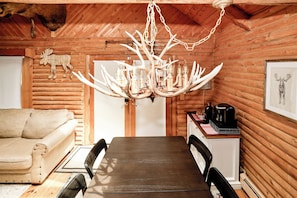 A rustic interior with a wooden dining table, chairs, a chandelier with antlers, a moose wall decoration, and a framed picture.