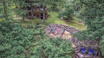 A rustic cabin surrounded by dense forest, with a stone fire pit and outdoor seating area.