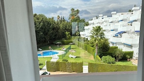 A view from a balcony with a swimming pool, neatly trimmed hedges, and a row of white buildings.