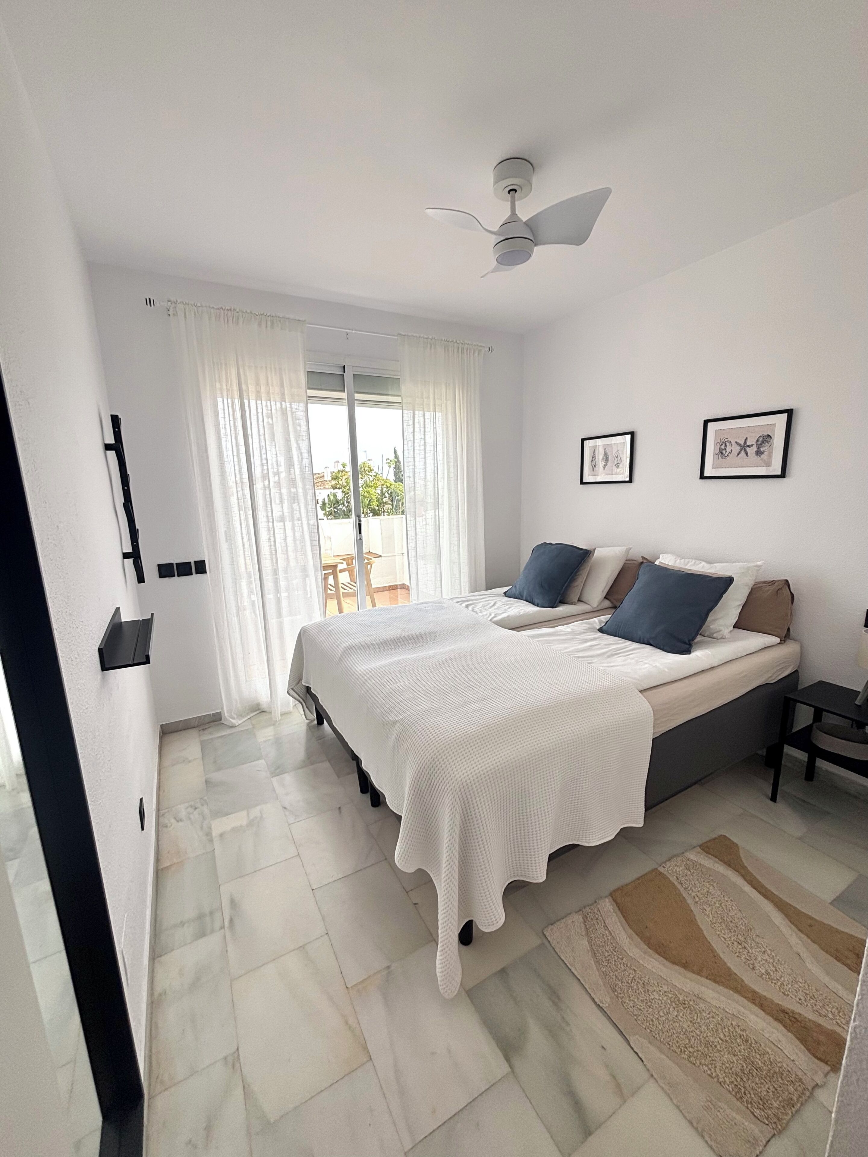 Un dormitorio con cama, ventilador de techo, ventana con cortinas y vistas al exterior.