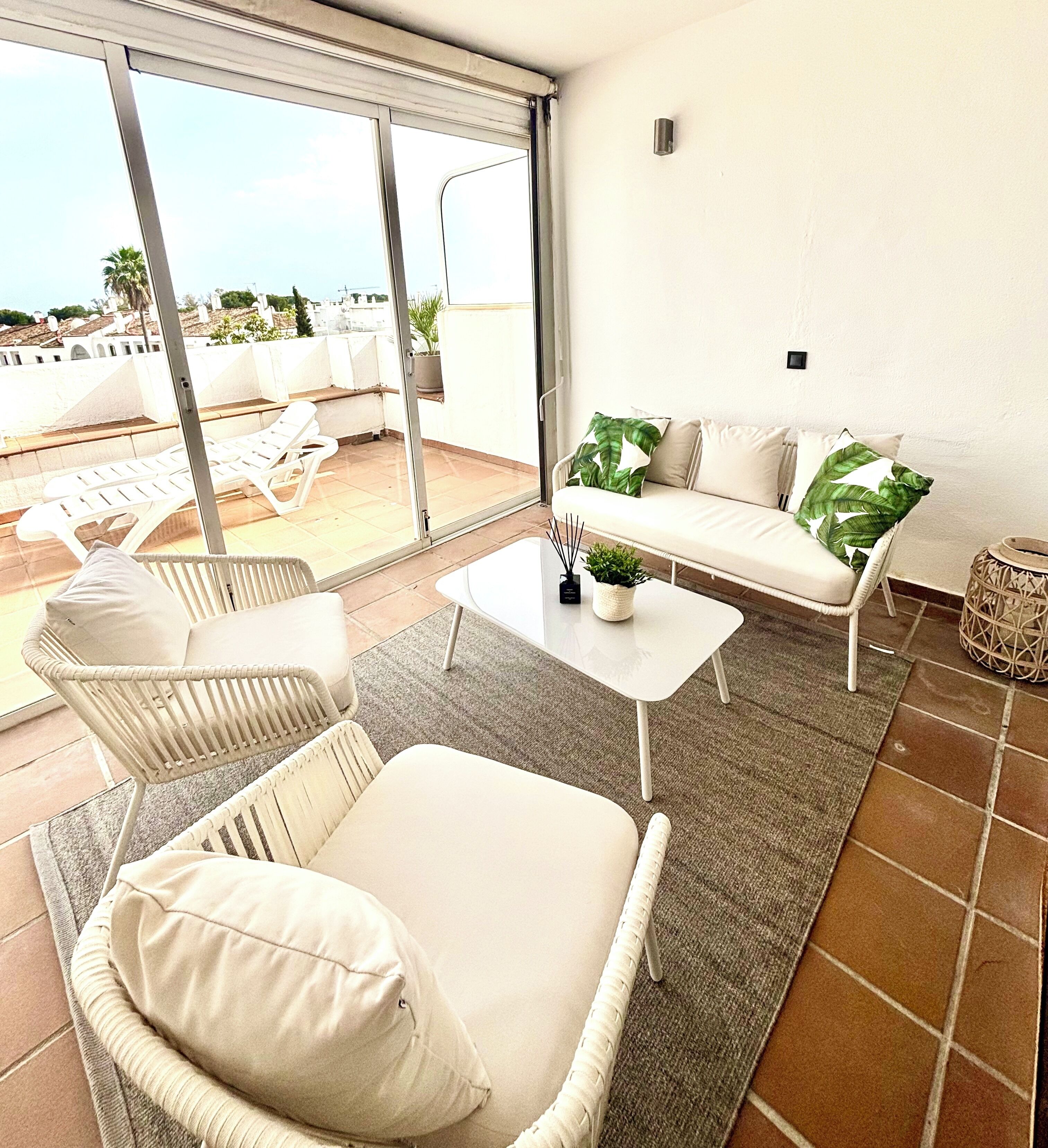 A balcony with white wicker furniture, a white sofa with green cushions, and a small table with a plant.