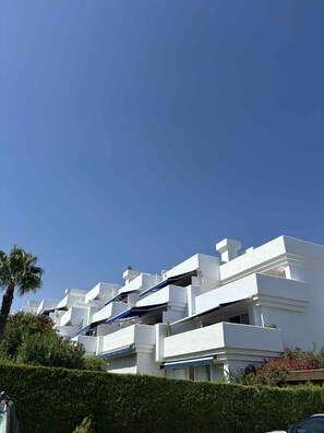 A modern white building with balconies, surrounded by greenery and a clear blue sky.