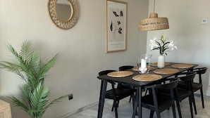 A dining area with a round table, black chairs, a woven placemat, a hanging light fixture, a framed artwork, and a potted plant.