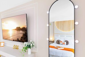 Ein Schlafzimmer mit einem Bett, einem Nachttisch, einem beleuchteten Spiegel, einem an der Wand befestigten Fernseher und einem Fenster mit Sonnenuntergangsblick.