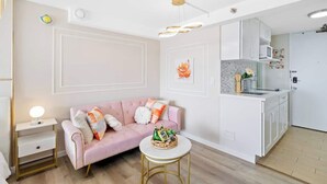 Ein modernes Wohnzimmer mit einem rosafarbenen Sofa, einem runden Couchtisch und einer Küchenzeile mit weißen Schränken und einer Mikrowelle.