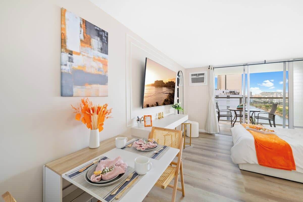 Habitación de hotel moderna con zona de comedor, cama con una manta naranja, televisor y balcón con vistas a la ciudad.