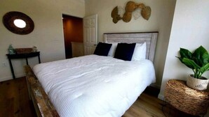 Quarto com cama, porta, objeto decorativo na parede e mesa de cabeceira em vime com uma planta.