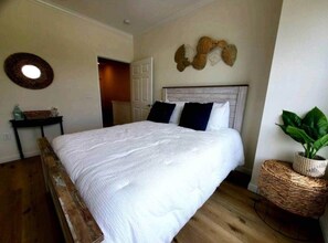 A bedroom with a bed, a door, a wall-mounted decorative item, and a wicker side table with a plant.