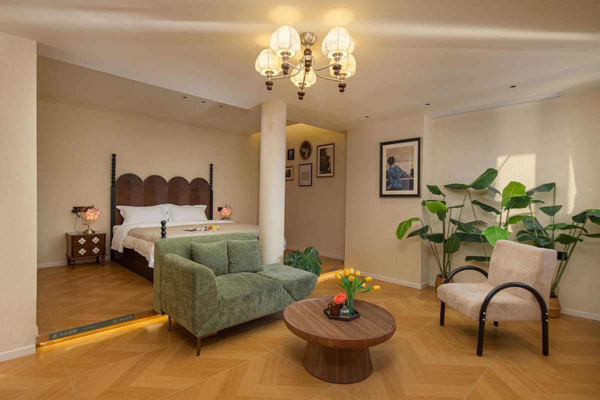 A bedroom with a bed, a green armchair, a wooden coffee table, and a chair with potted plants.