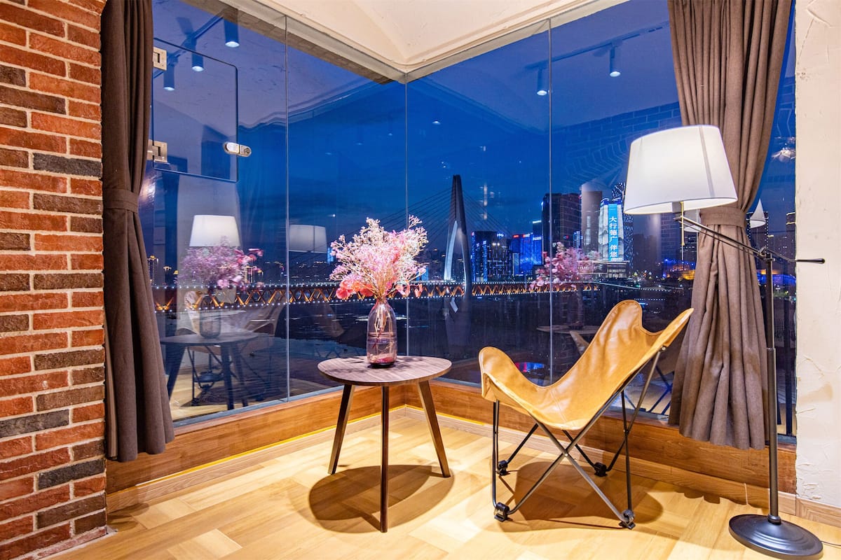 Une chambre moderne avec une grande fenêtre, une table ronde ornée d’un vase, un fauteuil en cuir et un lampadaire.