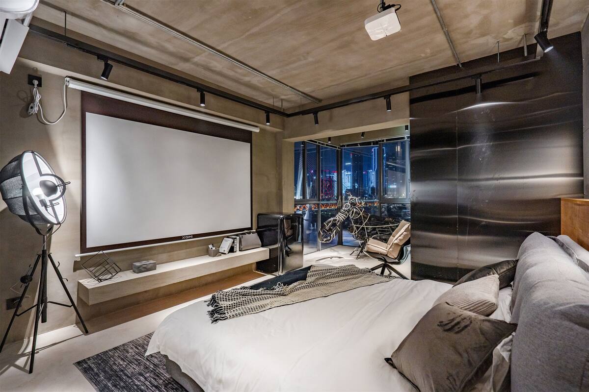 Une chambre moderne dotée d’un grand lit, d’un écran de projection et offrant une vue sur la ville.