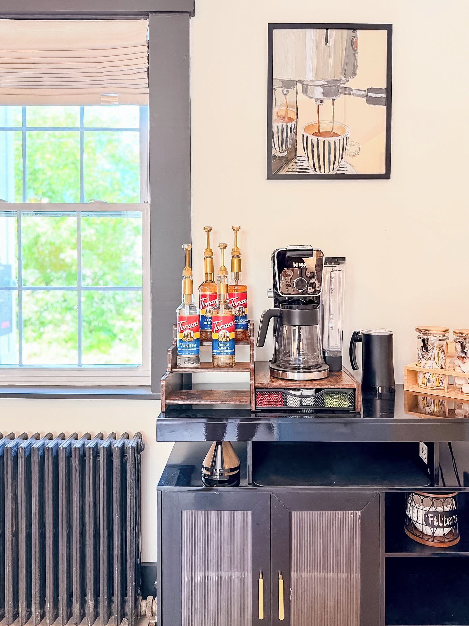 A kitchen area with a coffee machine, Torani syrup bottles, a radiator, and a framed artwork on the wall.