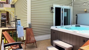 A hot tub on a deck with wooden chairs and a table, a door to a covered area, and string lights. - Downtown - Hot Tub, Speakeasy, Grill, Game Room, Fireplace (Ludlow)